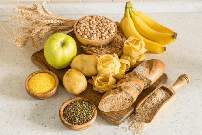 Divers féculents naturels comme pommes de terre, pâtes, pain, riz, pois chiches, bananes et pommes disposés sur une planche en bois.